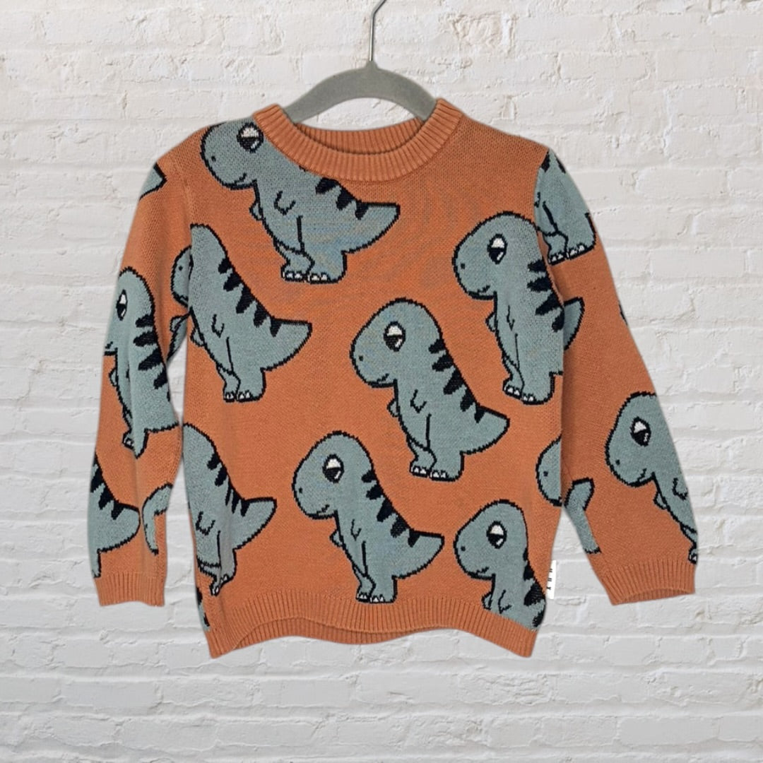 Orange knit sweater with grey dinosaur print, displayed on a hanger against a white brick background.