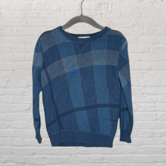 Blue plaid sweater with a round neckline hanging on a black hanger against a white brick wall.