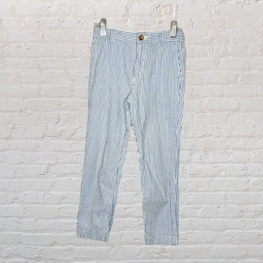 Blue and white striped seersucker pants with button closure and belt loops, hanging against a white brick wall.