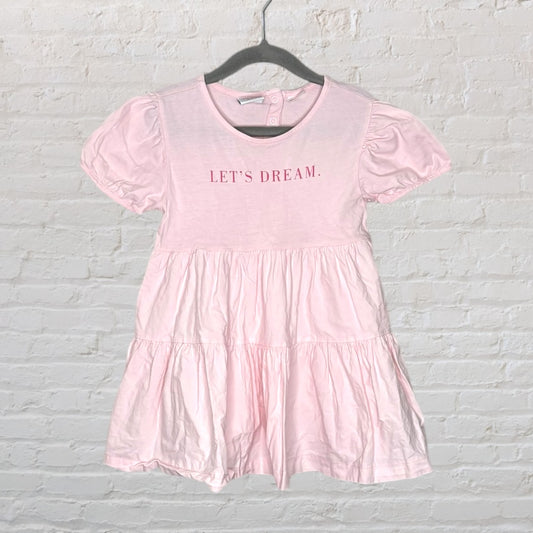 Light pink tiered dress with puff sleeves and 'Let's Dream' text on the front, hanging on a hanger.