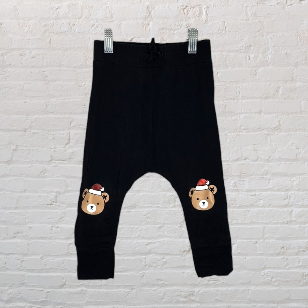 Black jogger pants with Christmas bear prints on knees, displayed against a white brick wall.