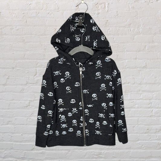 Black hoodie with white skull and crossbones pattern, hung on a hanger against a brick wall.