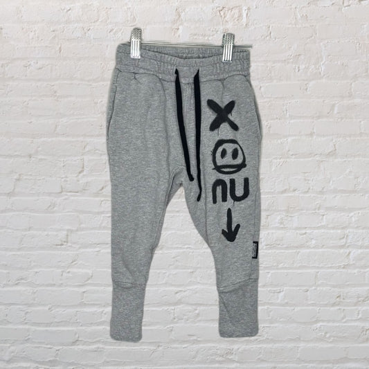 Grey NUNUNU sweatpants with black graphic designs, featuring an adjustable drawstring waist, hanging on a clip hanger.