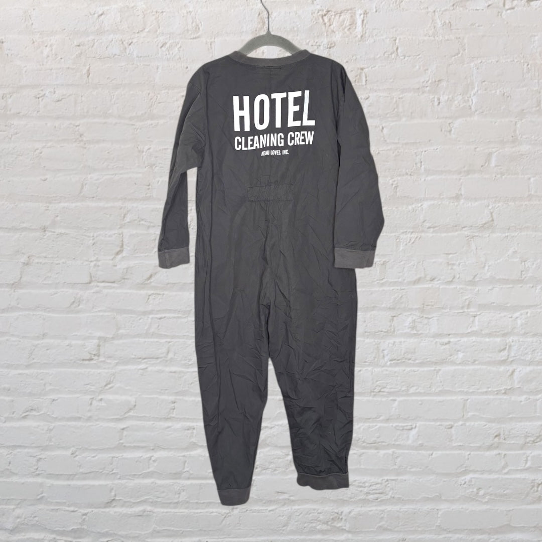 Dark grey coveralls with 'Hotel Cleaning Crew' printed on the back, displayed on a hanger against a white brick wall.
