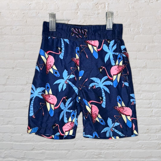 Navy blue swim shorts with pink flamingos and surfboard print, hanging against a white brick wall.