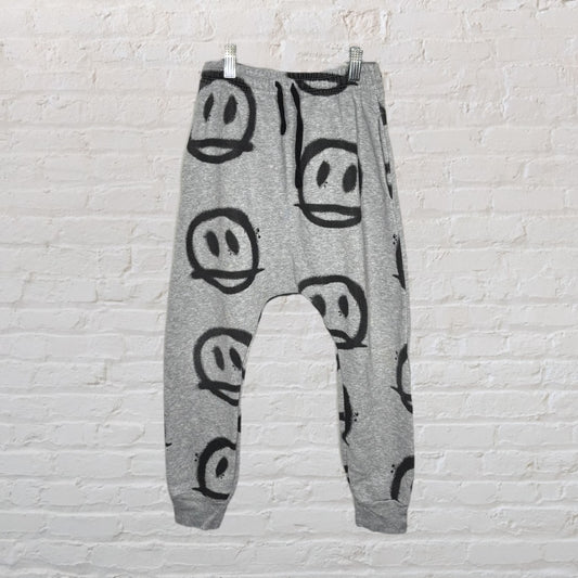 Grey jogger pants with large black smiley faces printed throughout, hanging against a white brick wall.