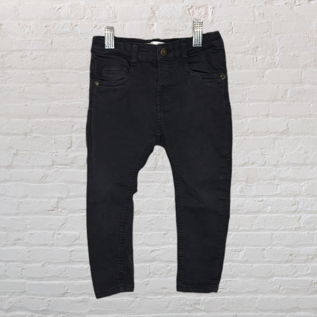 Children's black skinny jeans with front pockets and button closure, hanging against a brick wall.