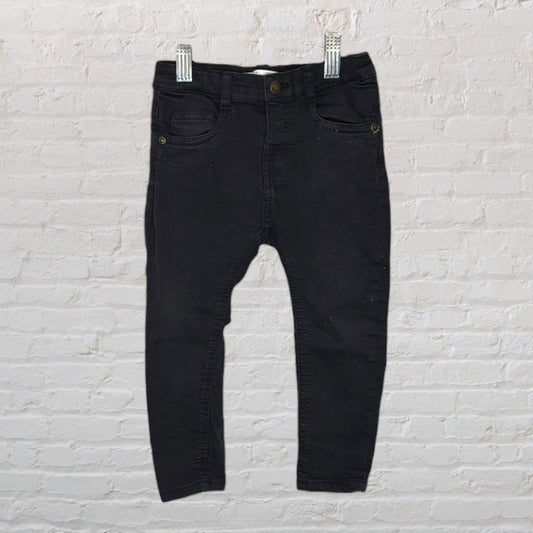 Children's black skinny jeans with front pockets and button closure, hanging against a brick wall.