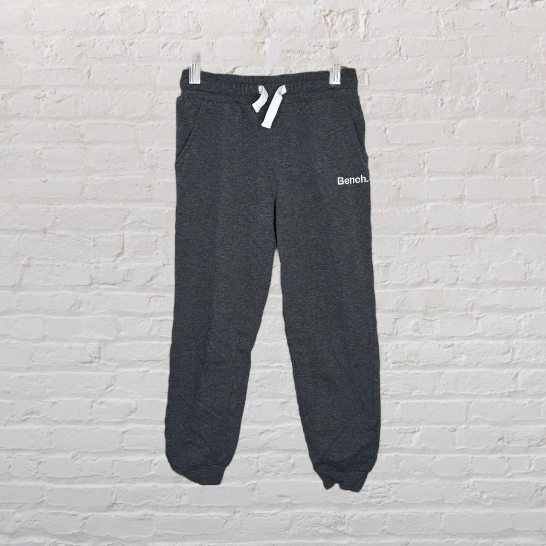 Dark grey Bench sweatpants with a drawstring waist and ribbed cuffs, hanging against a brick wall.