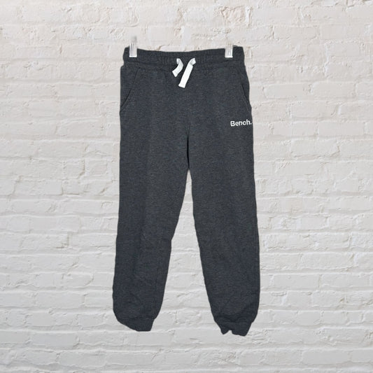 Dark grey Bench sweatpants with a drawstring waist and ribbed cuffs, hanging against a brick wall.