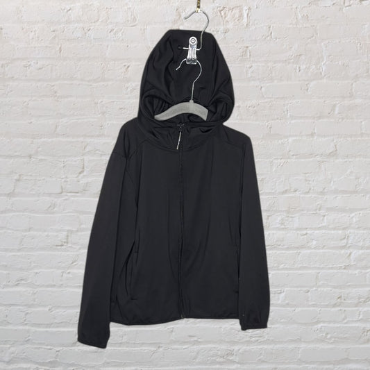 A plain black zip-up hoodie with a hood, hanging against a white brick wall.
