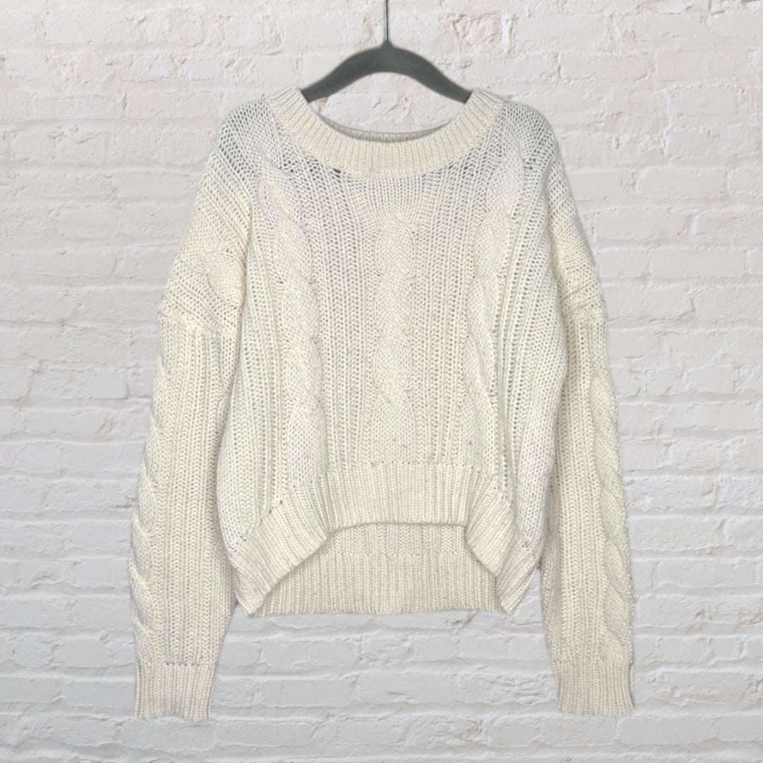 Cream cable knit sweater with a chunky design and ribbed trim, hanging on a hanger against a white brick wall.