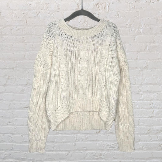 Cream cable knit sweater with a chunky design and ribbed trim, hanging on a hanger against a white brick wall.