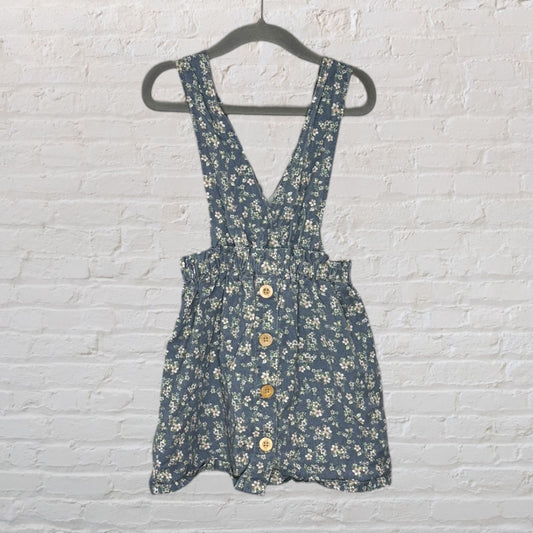 Blue floral print pinafore dress with wooden buttons, featuring adjustable straps and a gathered waist, hanging on a black hanger.