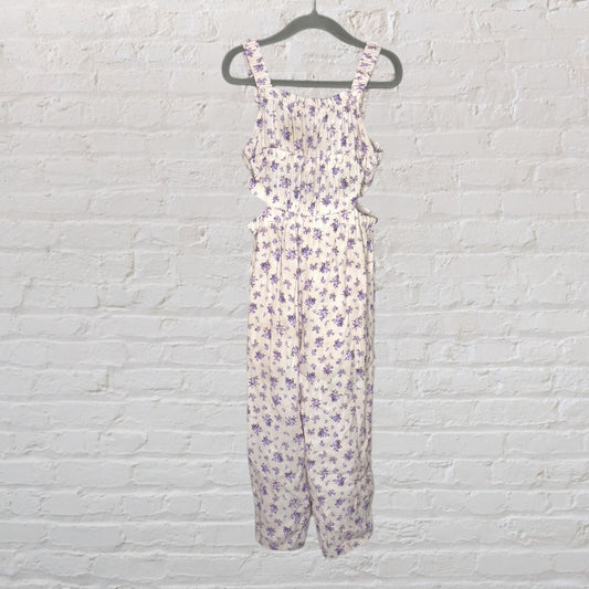 Cream floral print sleeveless jumpsuit with purple flowers hanging on a dark hanger against a white brick wall.