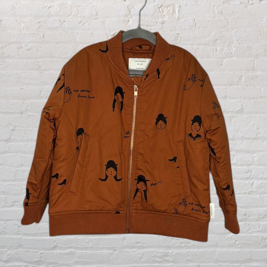 Brown bomber jacket with unique black graphics and text, displayed on a hanger against a white brick wall.