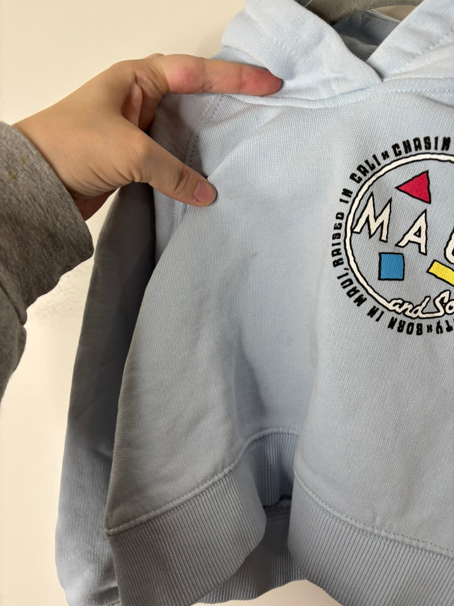 Light blue hoodie with a back graphic featuring 'Maui' and colorful geometric shapes, hanging against a white brick wall.