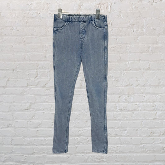 Light blue denim jeggings with elastic waistband hanging on a hanger against a white brick wall.