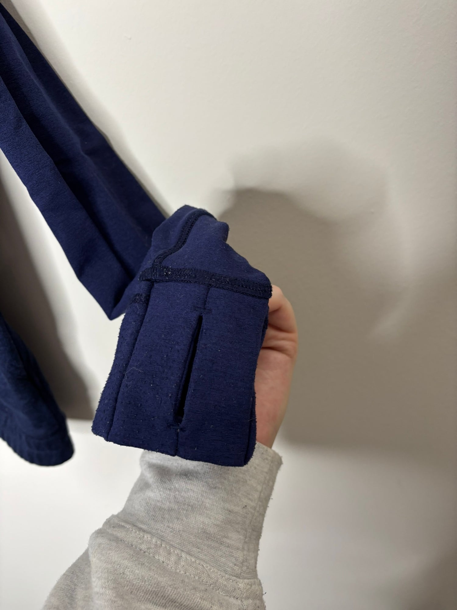 Navy blue full-zip jacket with a high neck and front pockets, hanging on a hanger against a white brick wall.
