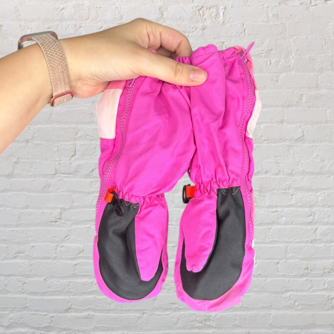 Kombi mittens in various shades of pink with logo, held by a hand against a brick background.