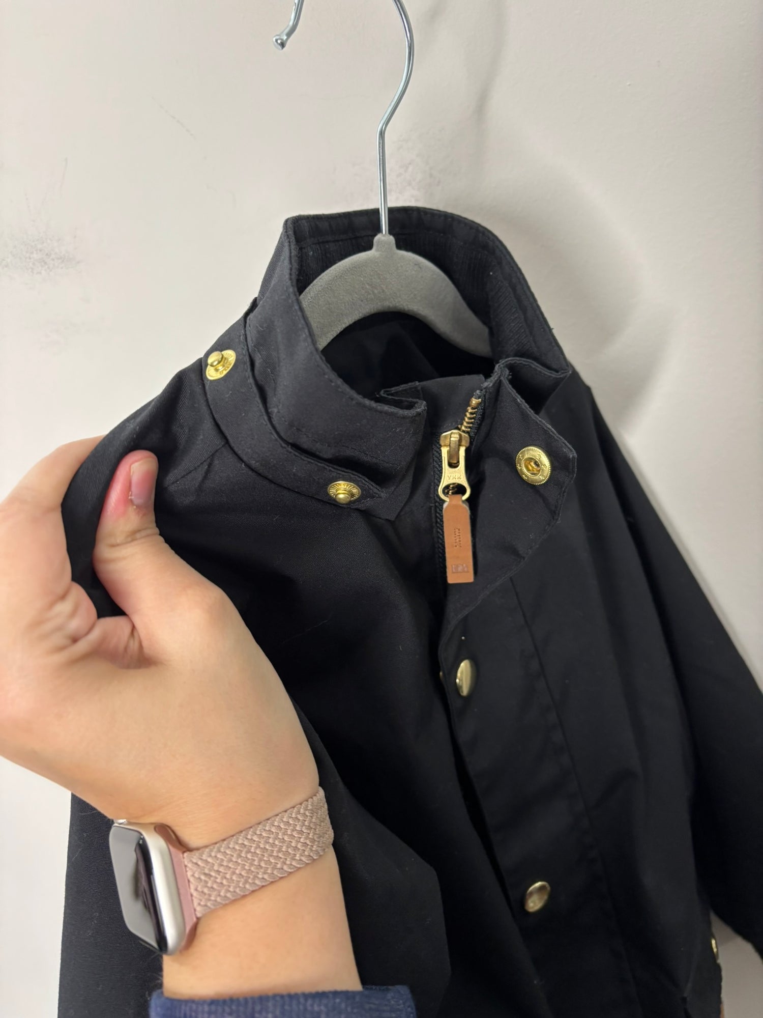 Kids black hooded parka jacket with gold buttons and front pockets, hanging on a hanger against a brick wall.