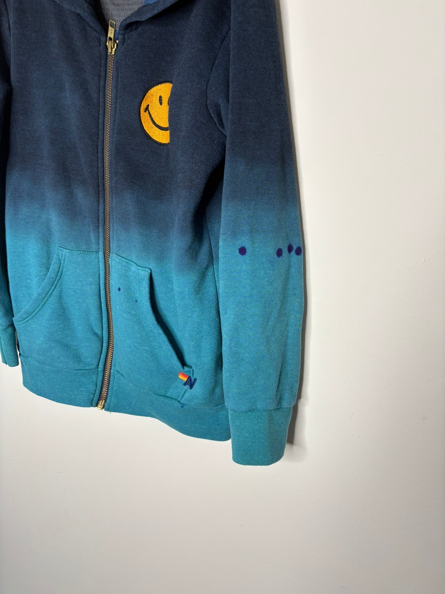 Navy and teal gradient zip-up hoodie with a smiley face patch and pockets, hanging on a white brick wall.