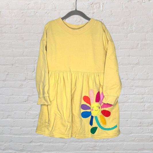 Yellow long sleeve dress with a colorful flower patch on the lower front, hung on a hanger against a white brick wall.