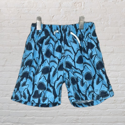 Blue swim shorts with a black shark pattern hanging by clips against a white brick wall.