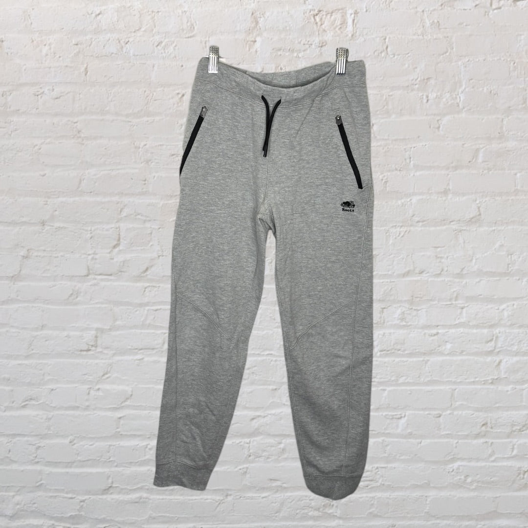 Grey Roots athletic jogger pants with black zipper pockets and drawstring, hanging against a white brick wall.
