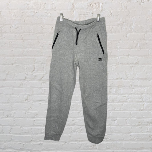 Grey Roots athletic jogger pants with black zipper pockets and drawstring, hanging against a white brick wall.