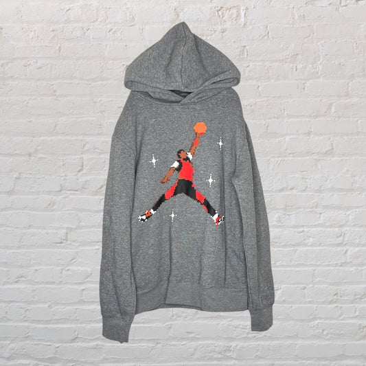 Gray hoodie featuring a pixel art basketball player graphic, hanging against a white brick wall.