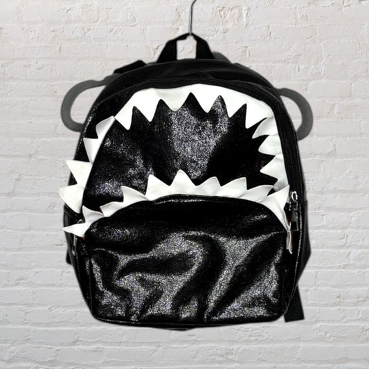 Black glittery shark-themed backpack with white jagged teeth design, hanging against a white brick wall.