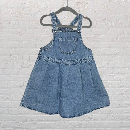 Blue denim overall dress with front pocket and pleated skirt, hanging on a black hanger.