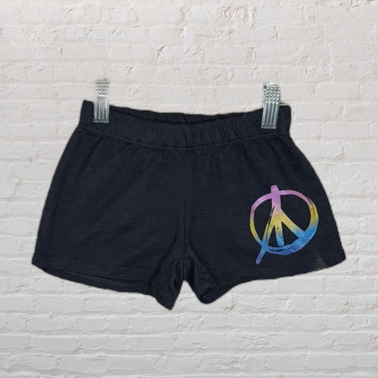 Black shorts with a multicolor peace sign graphic on the front, displayed on a hanger against a white brick background.