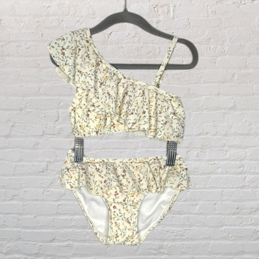 Women's cream floral one-shoulder bikini with ruffles on a hanger against a white brick wall.