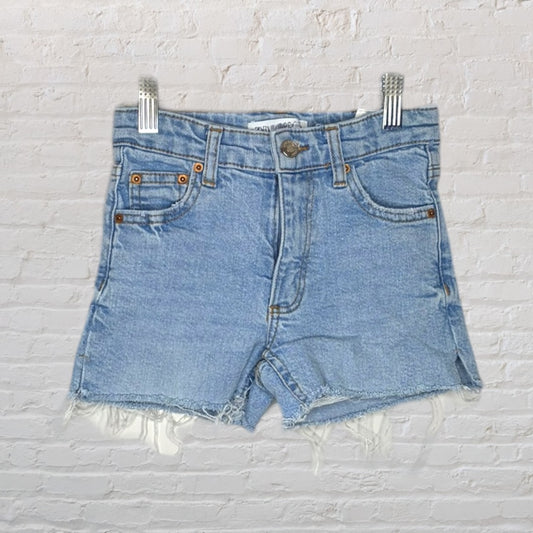 Light blue high-waisted denim shorts with frayed hems hanging against a white brick background.