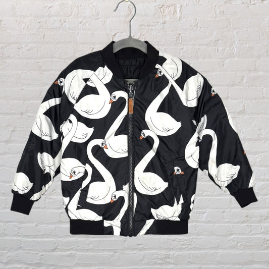 Unisex black bomber jacket featuring a white swan pattern, displayed on a hanger against a brick wall.