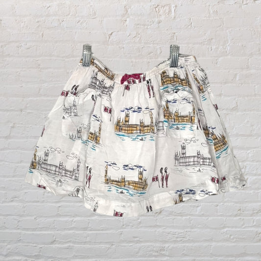 Women's white skirt with a scenic print, including landmarks and people, displayed on a hanger against a brick wall.