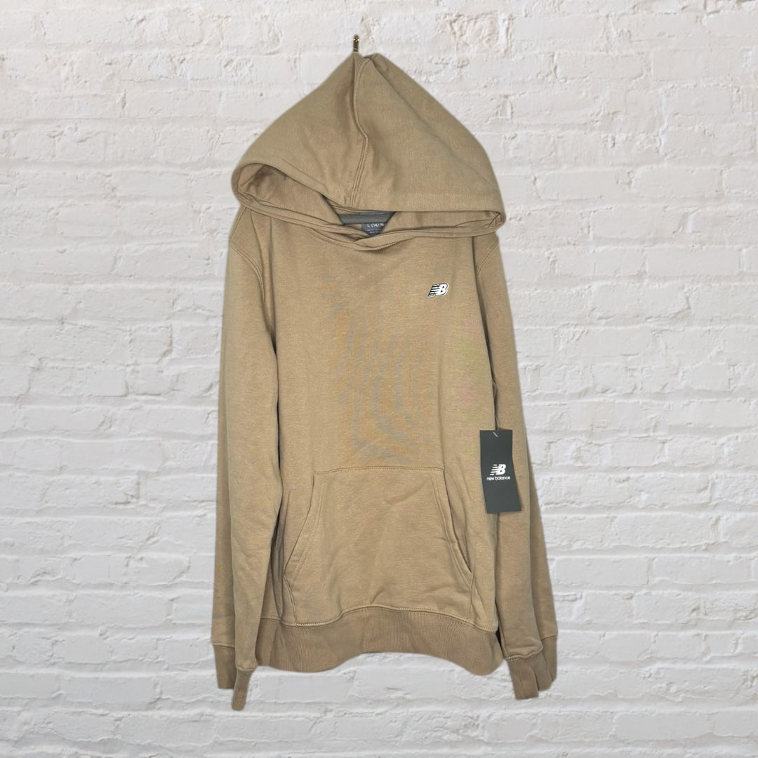 Beige New Balance hoodie with front pocket and logo, new with tags, hanging against a white brick wall.