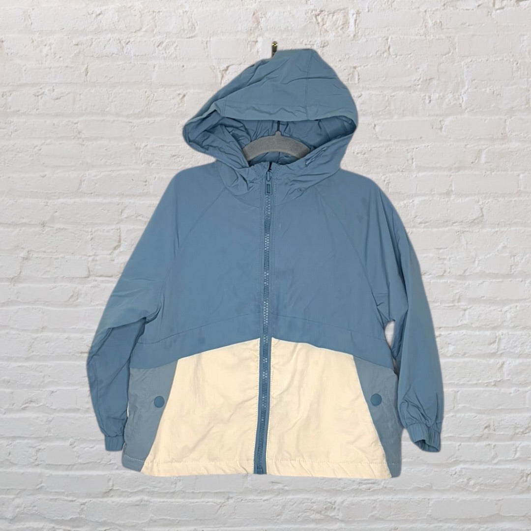 Blue and white hooded windbreaker jacket with front zipper and pockets, displayed on a hanger against a white brick wall.