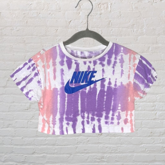 Purple and pink tie-dye Nike crop top with a blue logo, displayed on a hanger against a brick wall.