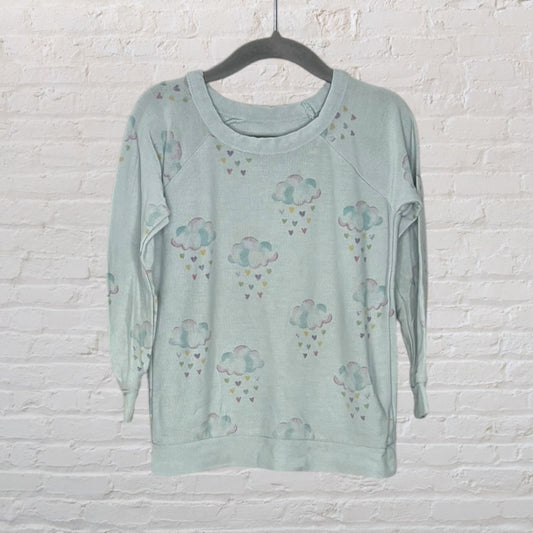 Light blue long sleeve top with a cloud and heart pattern, hanging against a white brick wall.