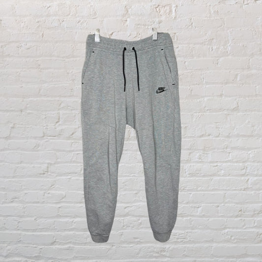 Grey Nike jogger pants with drawstring waistband and logo on left leg, hanging against a white brick wall.