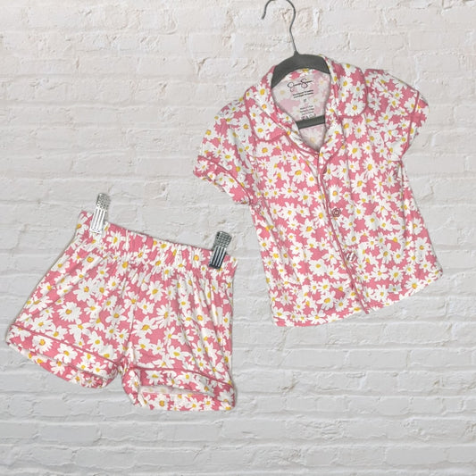 Pink floral short sleeve pajama set with button-up top and elastic waist shorts, hanging against a white brick wall.