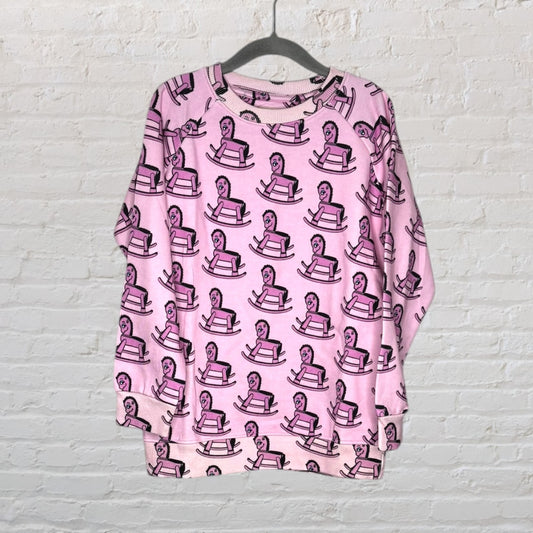 Pink sweatshirt featuring a playful all-over pattern of rocking horses, hanging against a brick wall.
