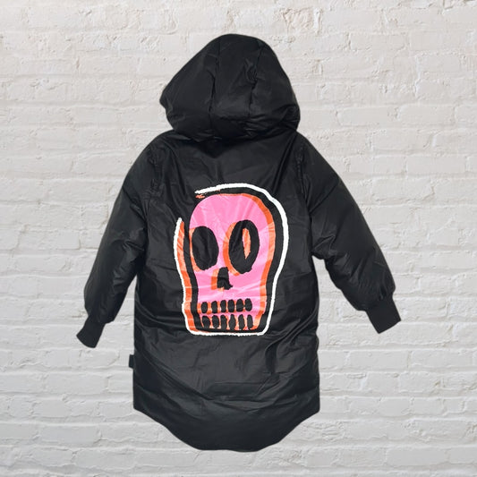 Black hooded puffer jacket featuring a large pink skull graphic on the back, hung against a brick wall background.