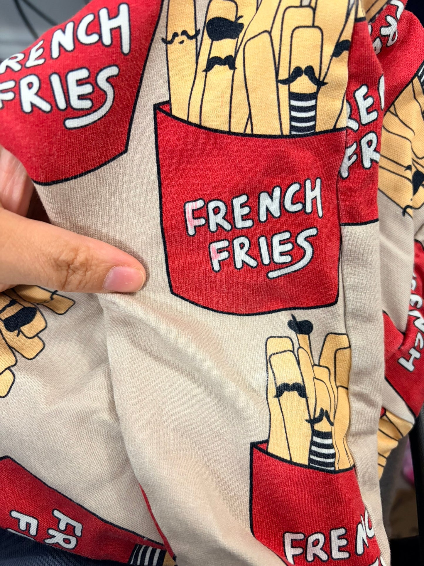 Beige jogger pants with a playful French fries graphic print, featuring a drawstring waist, hanging on clips against a white brick wall.