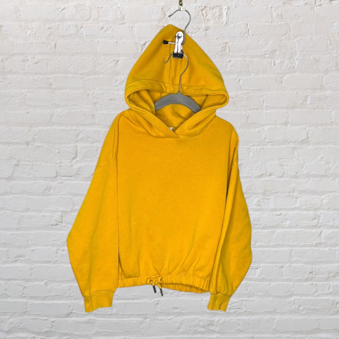 Bright yellow hoodie with drawstring hem and relaxed fit, displayed on a hanger against a white brick wall.