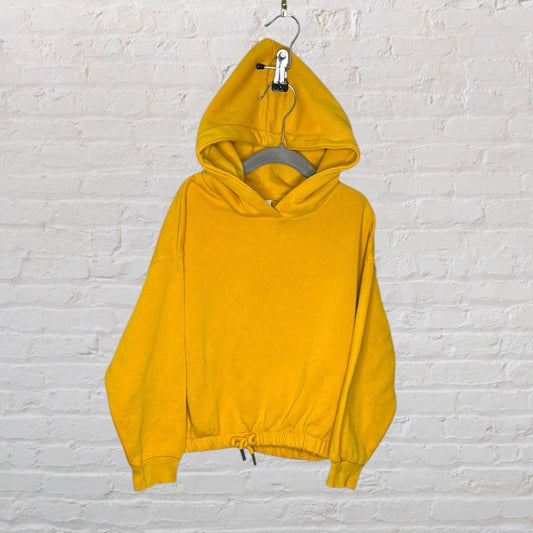 Bright yellow hoodie with drawstring hem and relaxed fit, displayed on a hanger against a white brick wall.