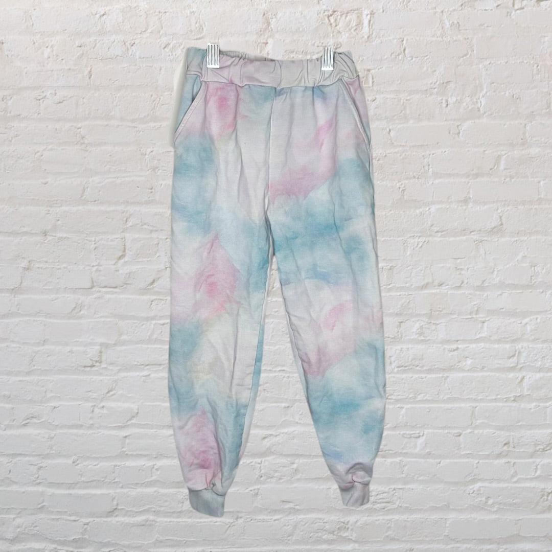 Unisex pastel tie-dye jogger pants in shades of pink, blue, and white, hanging on white clips against a white brick wall.